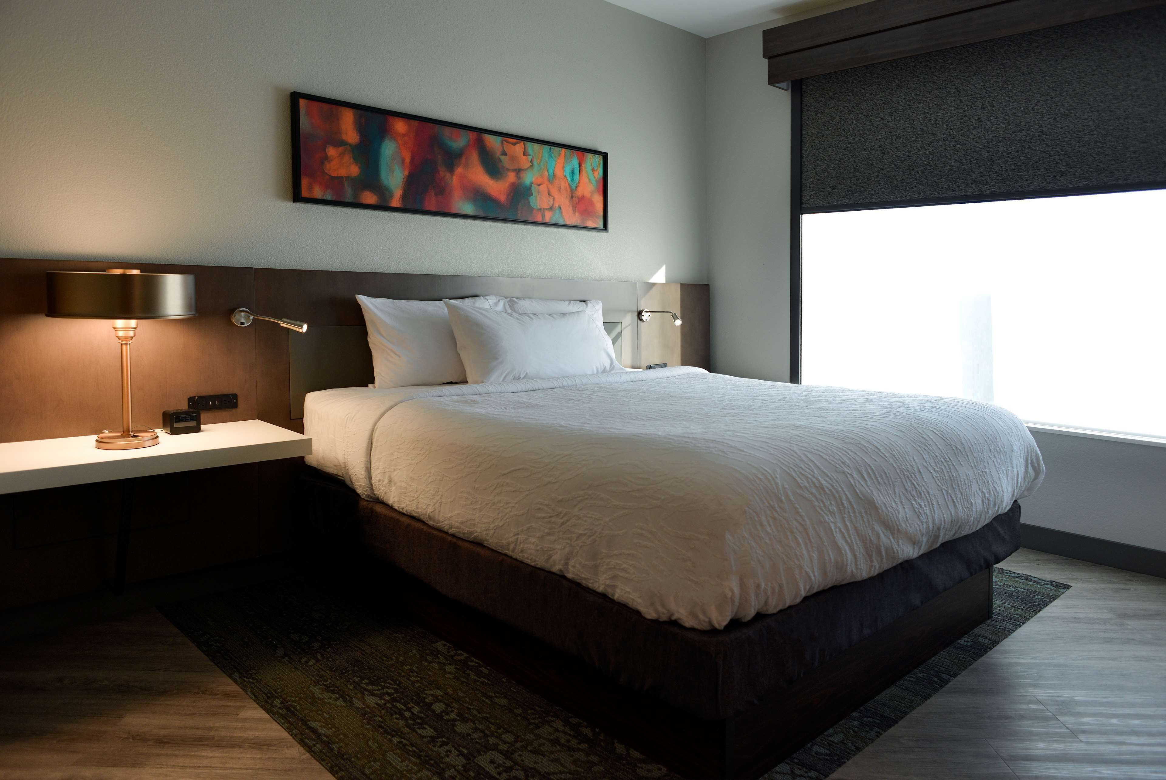 A modern hotel room with a large bed, a bedside table with a lamp, and a window with a view.