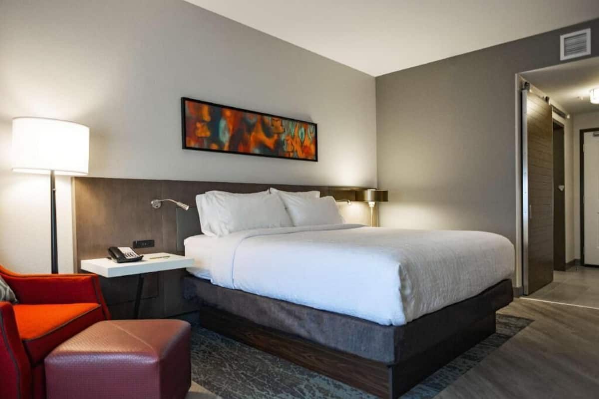 A hotel room with a bed, bedside table, lamp, and a red chair.