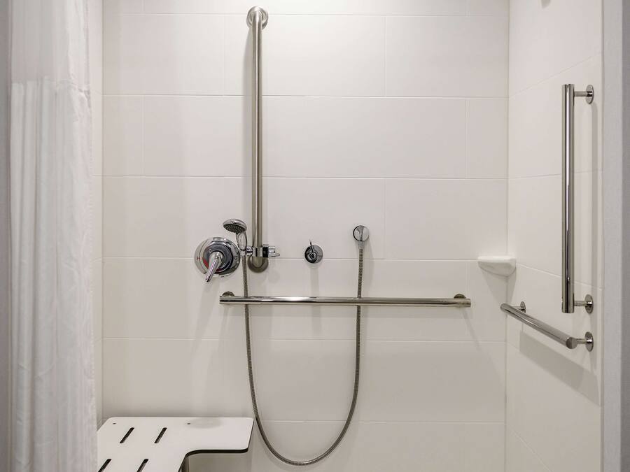 Um chuveiro acessível com azulejos brancos, chuveiro manual, barra de apoio e um pequeno banco.