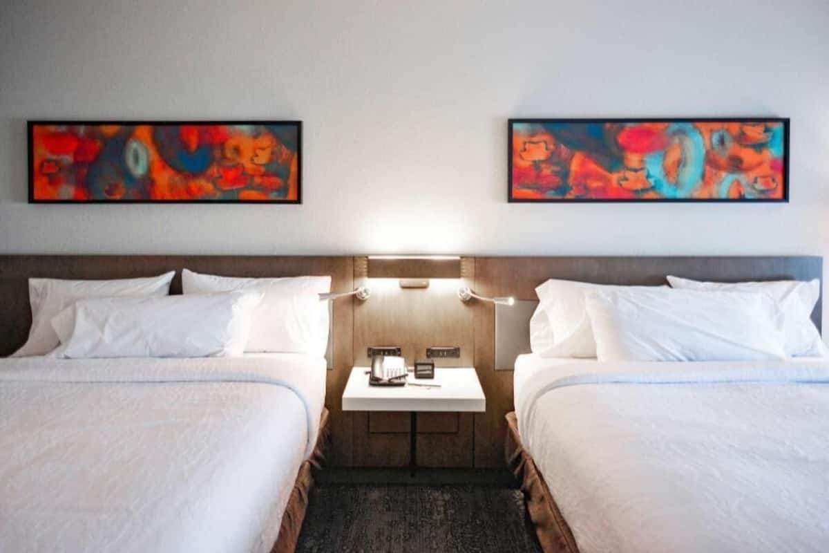 A hotel room with two beds, a small table with a lamp, and abstract paintings on the wall.