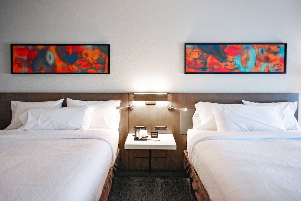 A hotel room with two beds, a small table with a phone and a lamp, and abstract paintings on the wall.