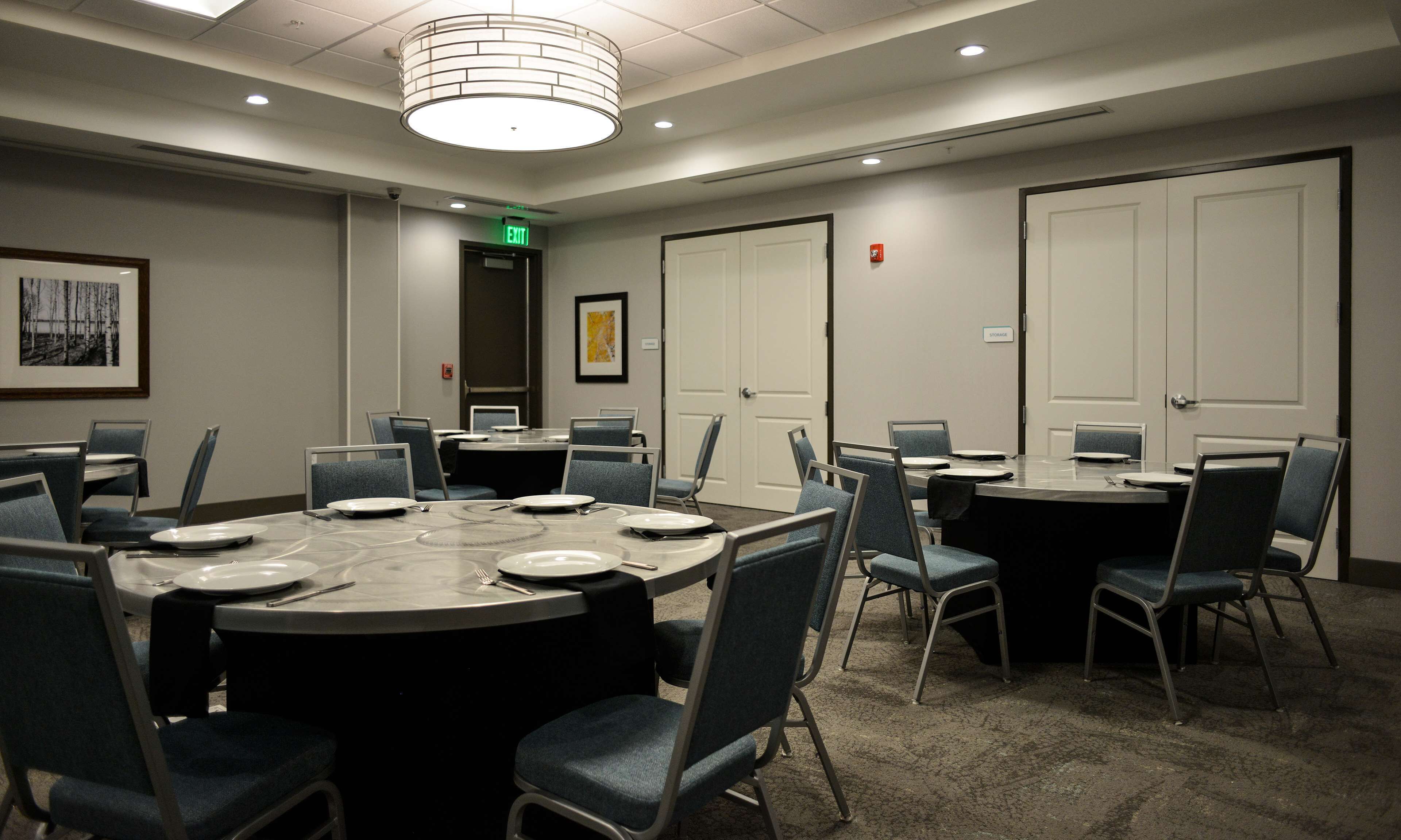A conference room with a large oval table, multiple chairs, a door with an exit sign, and a framed picture on the wall.