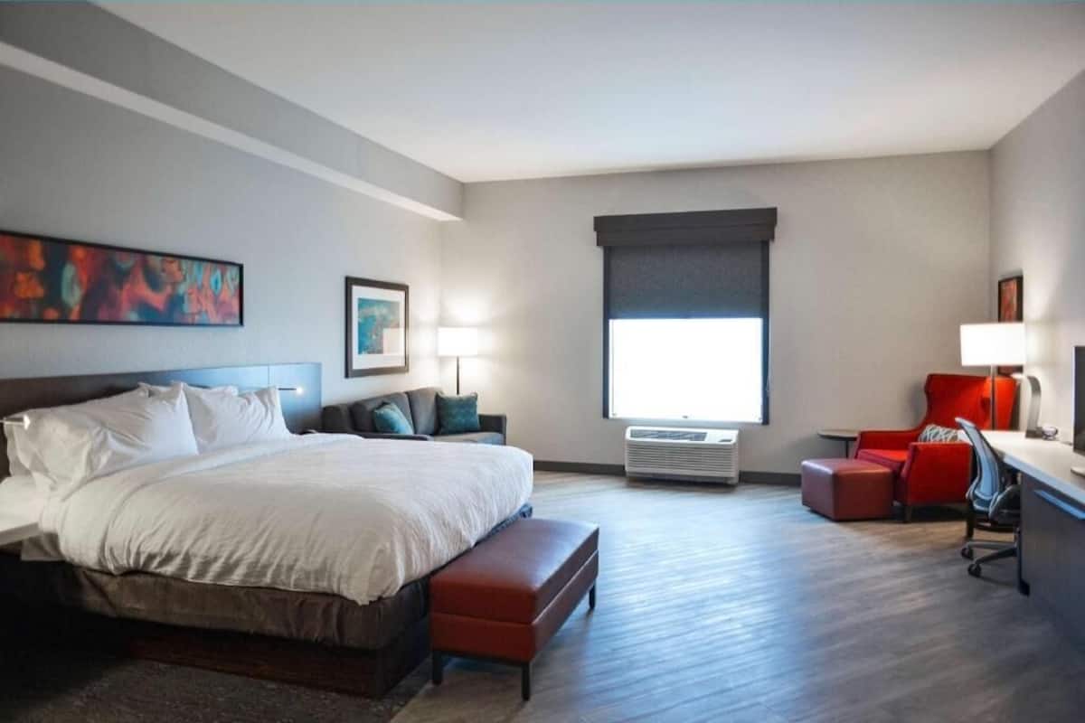 A hotel room with a bed, a desk, a chair, an air conditioning unit, and a window.