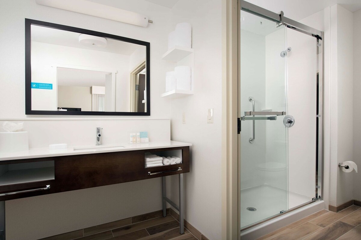 Un baño moderno con cabina de ducha de vidrio, un espejo grande y un mueble de tocador con detalles en madera oscura.