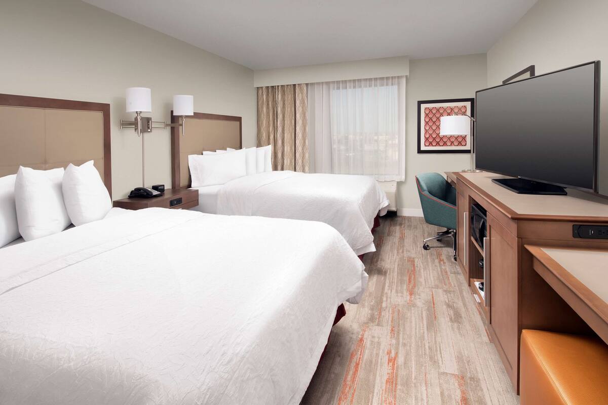 Habitación de hotel con dos camas, un escritorio, un televisor de pantalla plana, una silla y un ventanal con cortinas.