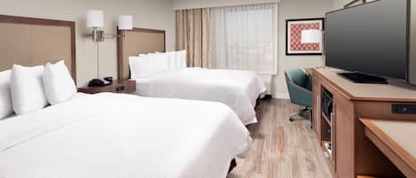 A hotel room with two beds, a desk with a flat-screen TV, a chair, and a large window with curtains.