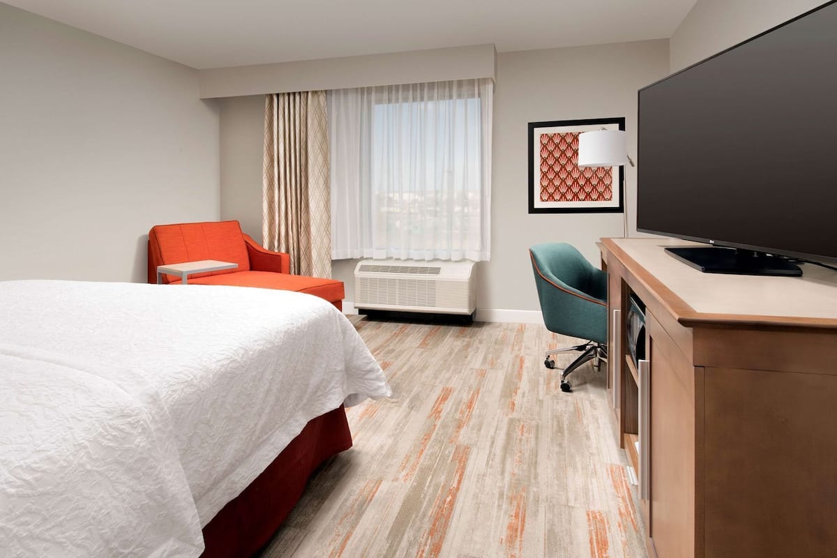 Una habitación de hotel con cama, escritorio con televisor, silla y ventana con cortinas.