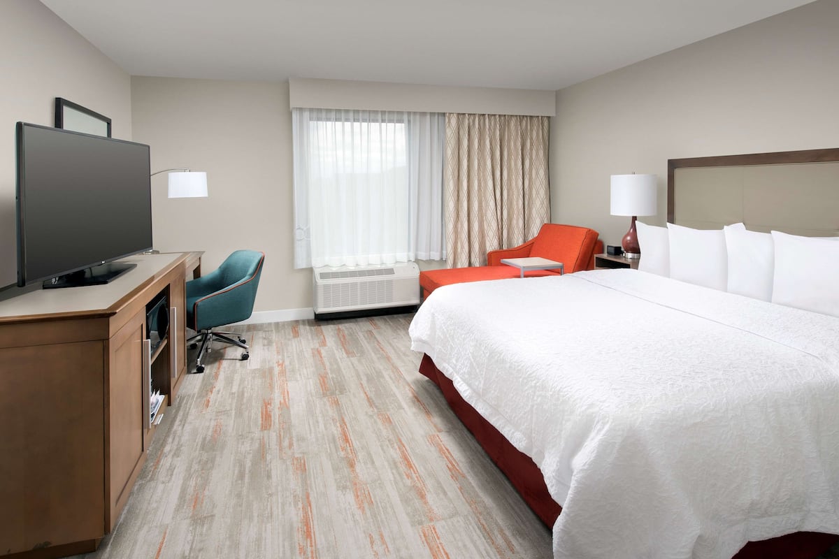Habitación de hotel con cama, escritorio con televisor, una silla, lámpara y ventana con cortinas.