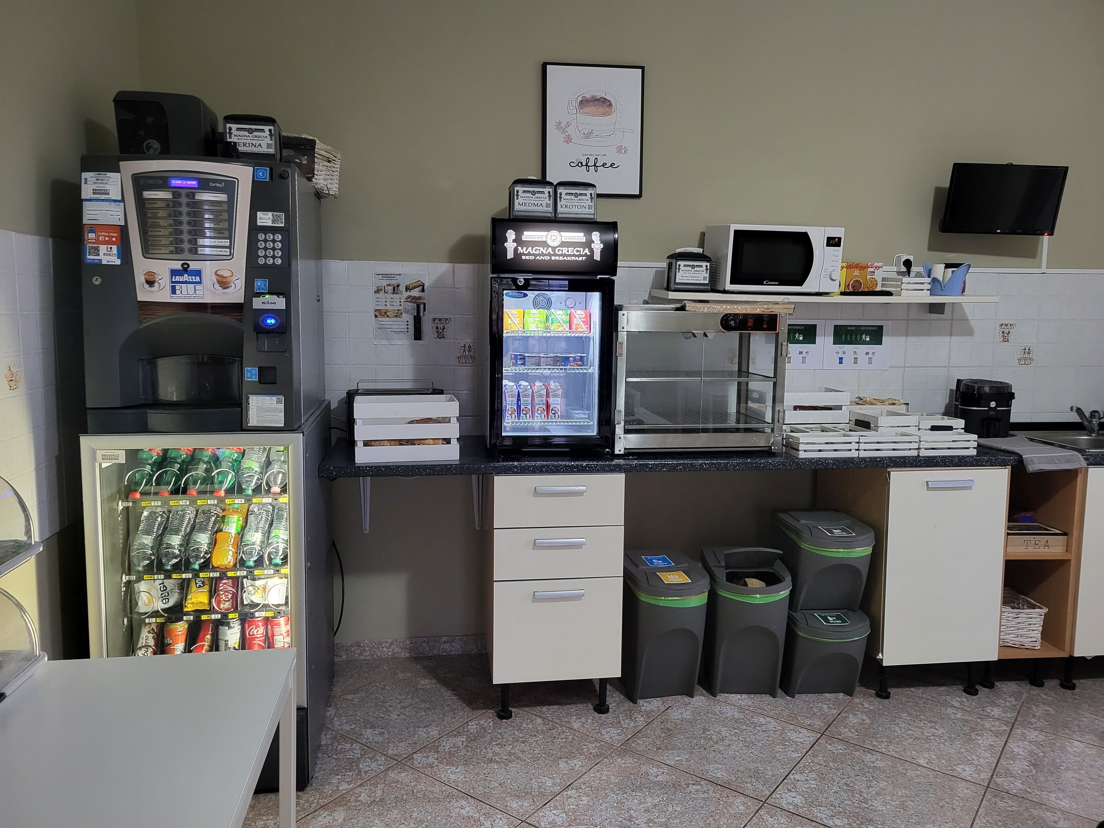Una zona de kitchenette con cafetera, microondas y refrigerador. Hay botes de basura y una máquina expendedora de bebidas.