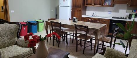 A compact kitchen with wooden cabinets, a dining table, and chairs. There is a small refrigerator, a sink, and a stove. A vase with red flowers is on a table.
