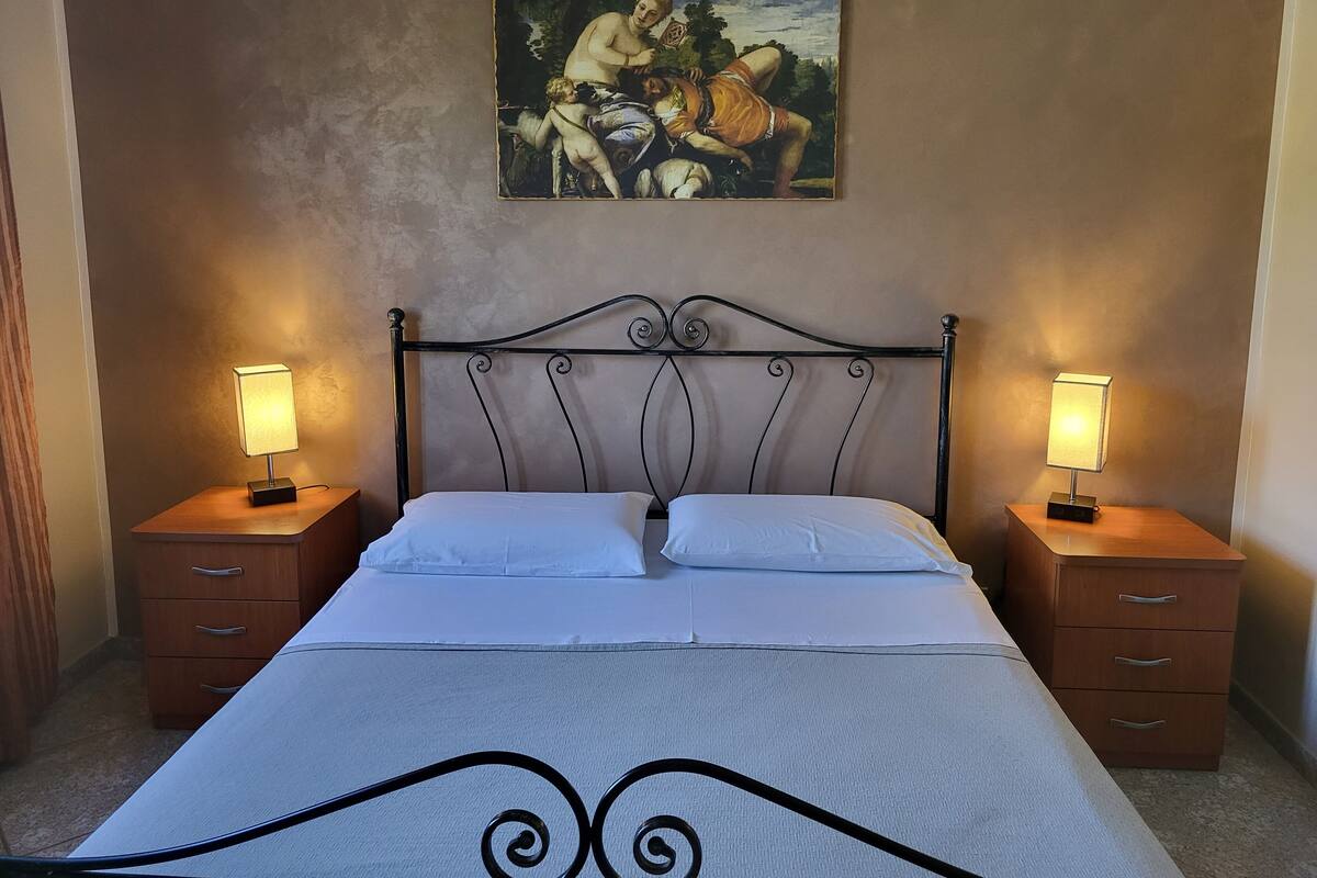 Ein Schlafzimmer mit einem Bett, zwei Nachttischen, einem Schminktisch, einem Bild an der Wand und einem verzierten Eiserne-Kopfteil.