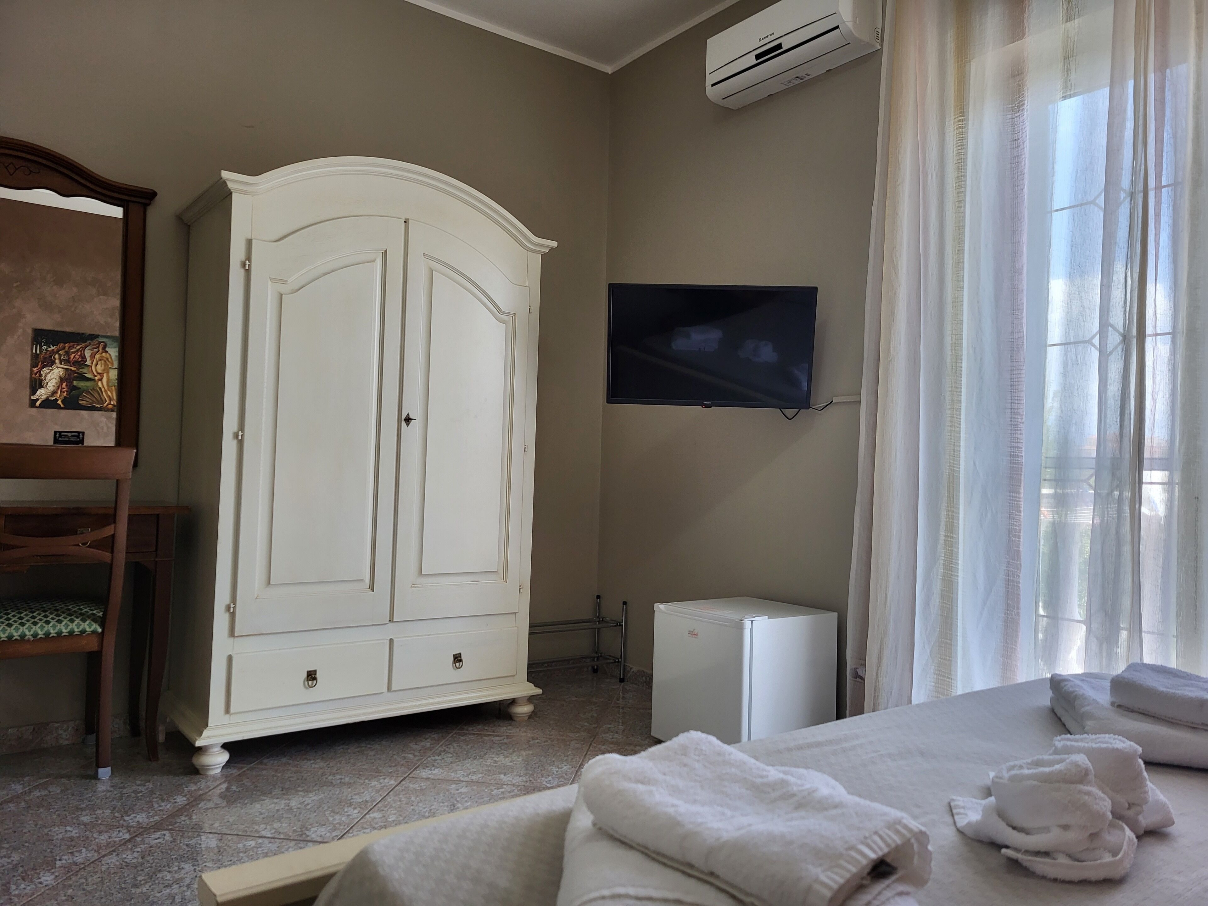 Ein Hotelzimmer mit weißem Kleiderschrank, einem Flachbildfernseher, einer Minikühlschrank und einem Bett mit ordentlich gefalteten Handtüchern.