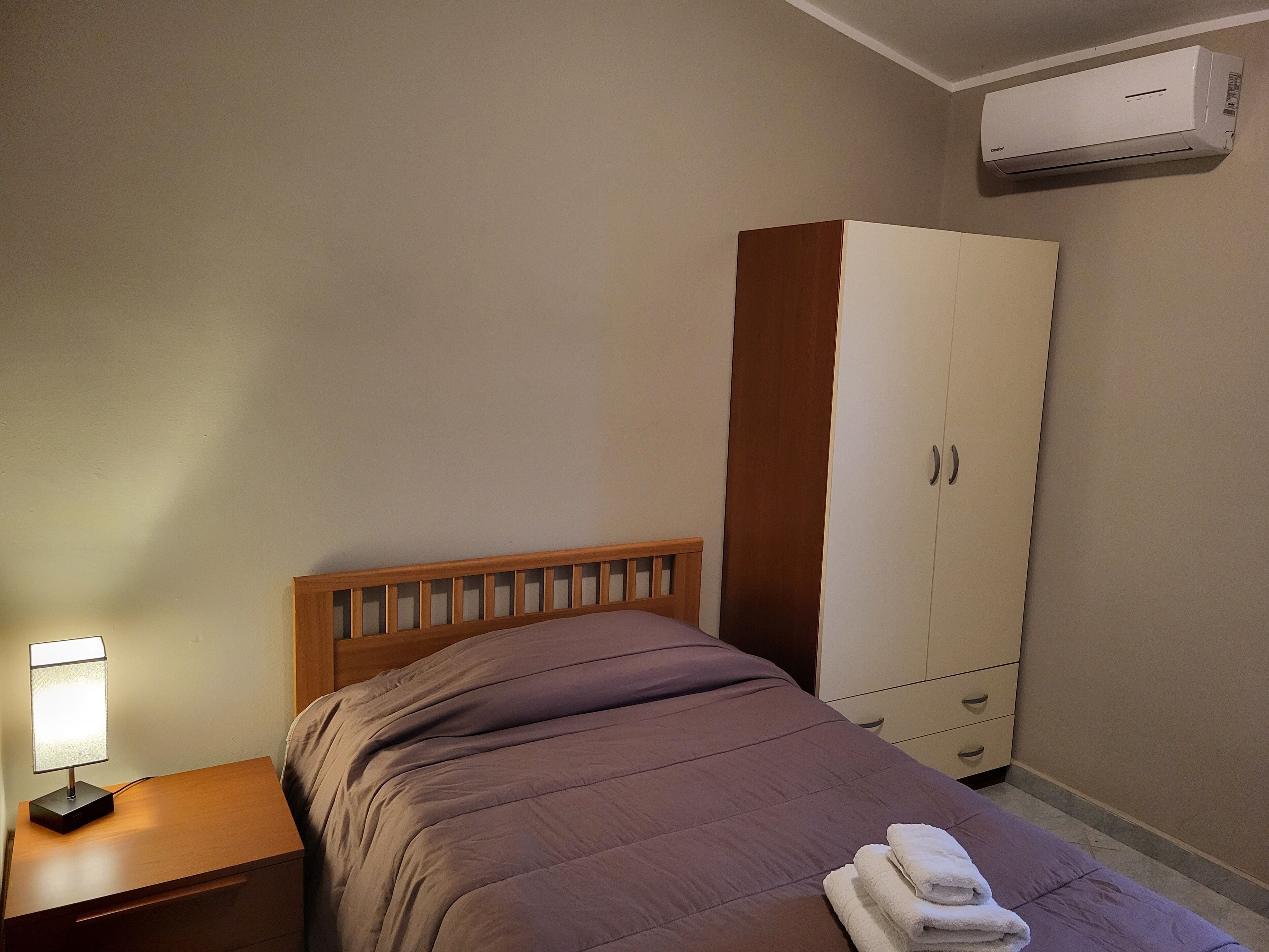Une chambre à coucher comprenant un lit, une table de chevet, une lampe, une armoire et un appareil de climatisation.