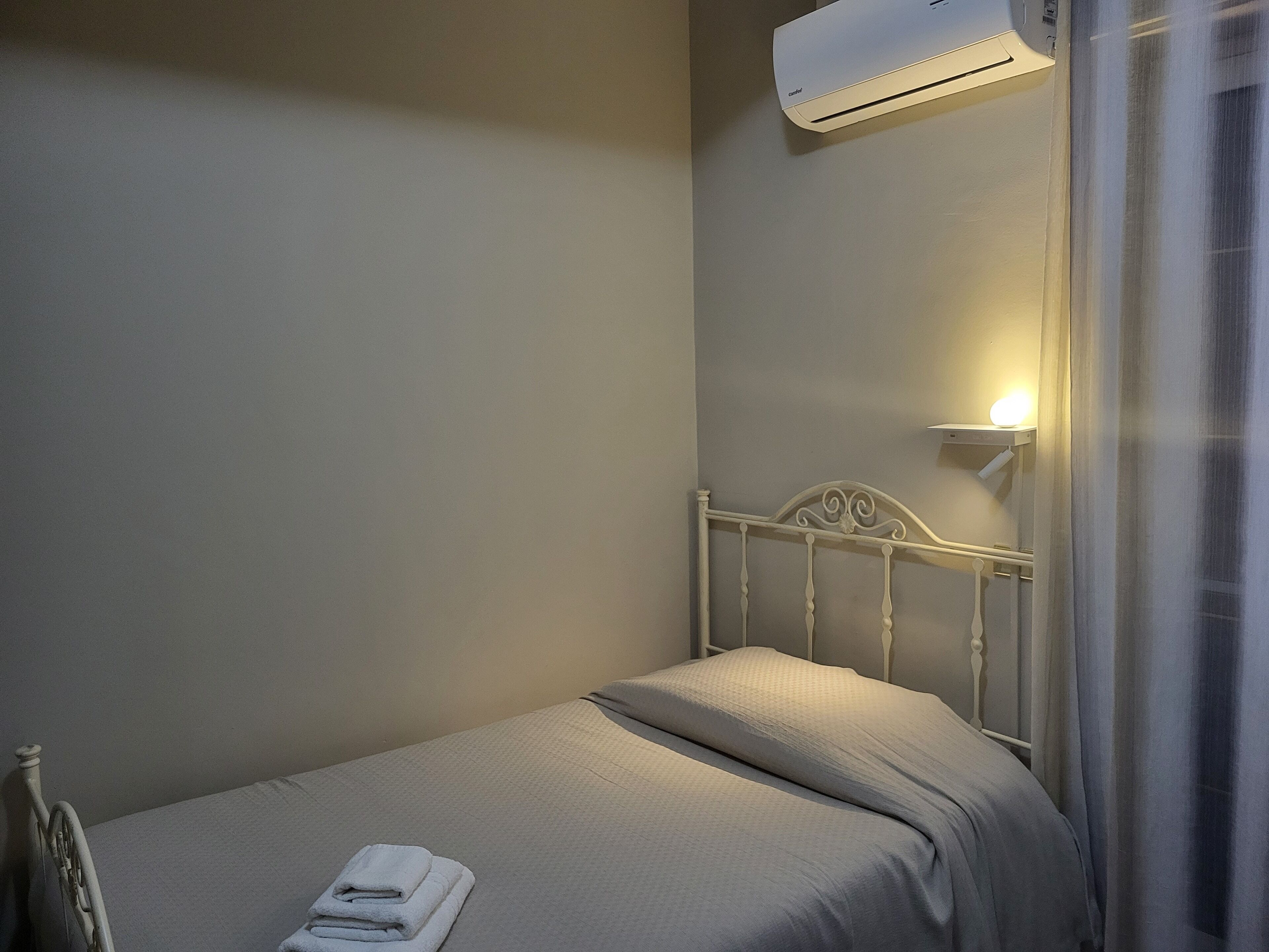 A single bed with a white headboard, a bedside table with a lamp, and an air conditioning unit mounted on the wall.