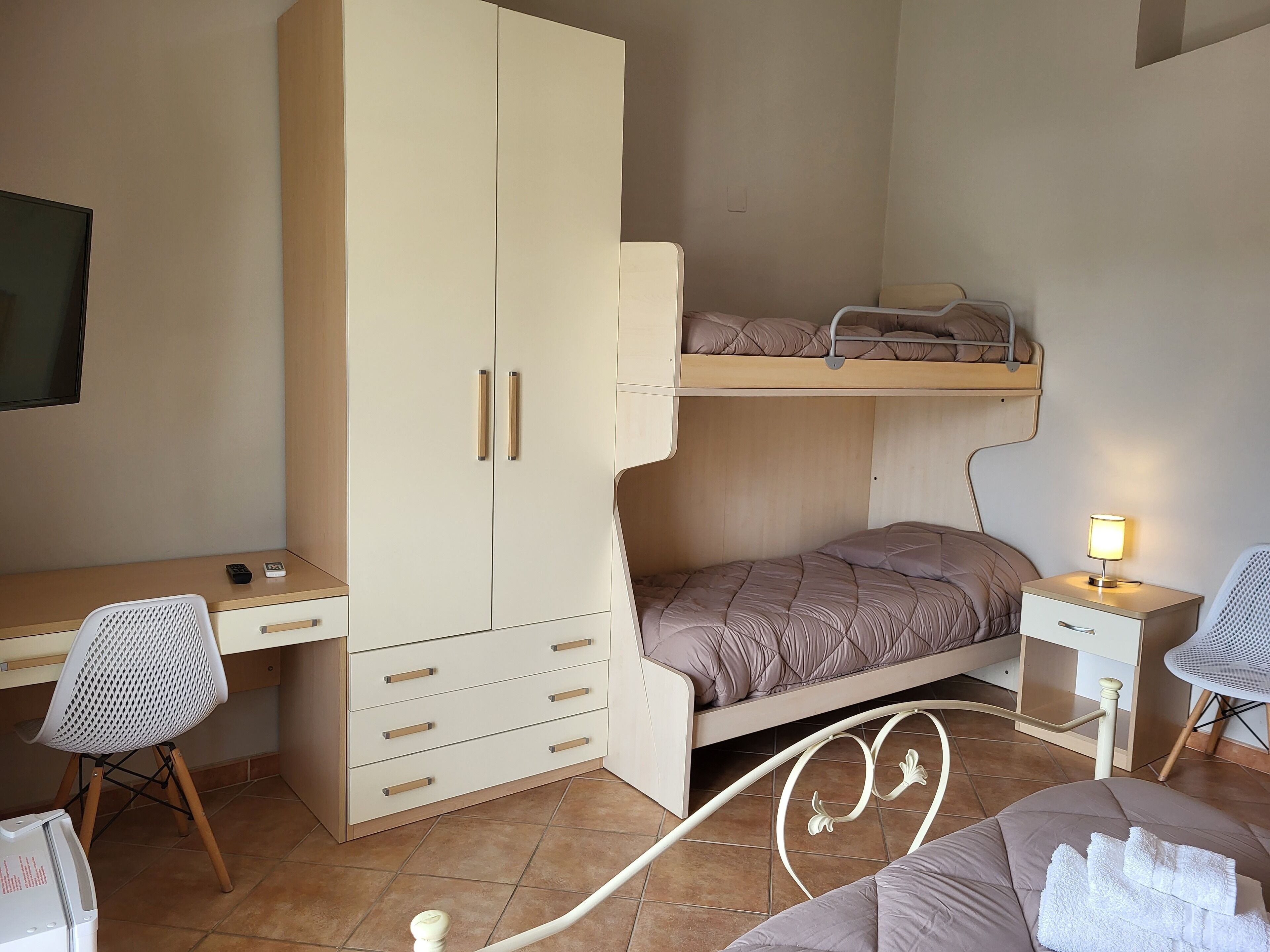 A compact bedroom with a bunk bed, a desk, a chair, and a bedside table with a lamp.