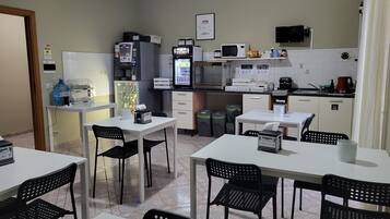 Une petite cuisine équipée d’un four à micro-ondes, d’un réfrigérateur et d’une machine à café. On y trouve deux tables avec des chaises, une poubelle et un distributeur d’eau.