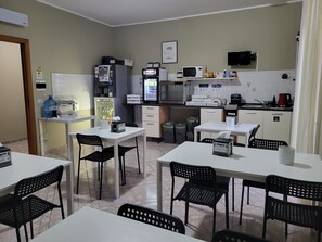 Une petite cuisine équipée d’un four à micro-ondes, d’un réfrigérateur et d’une machine à café. On y trouve deux tables avec des chaises, une poubelle et un distributeur d’eau.