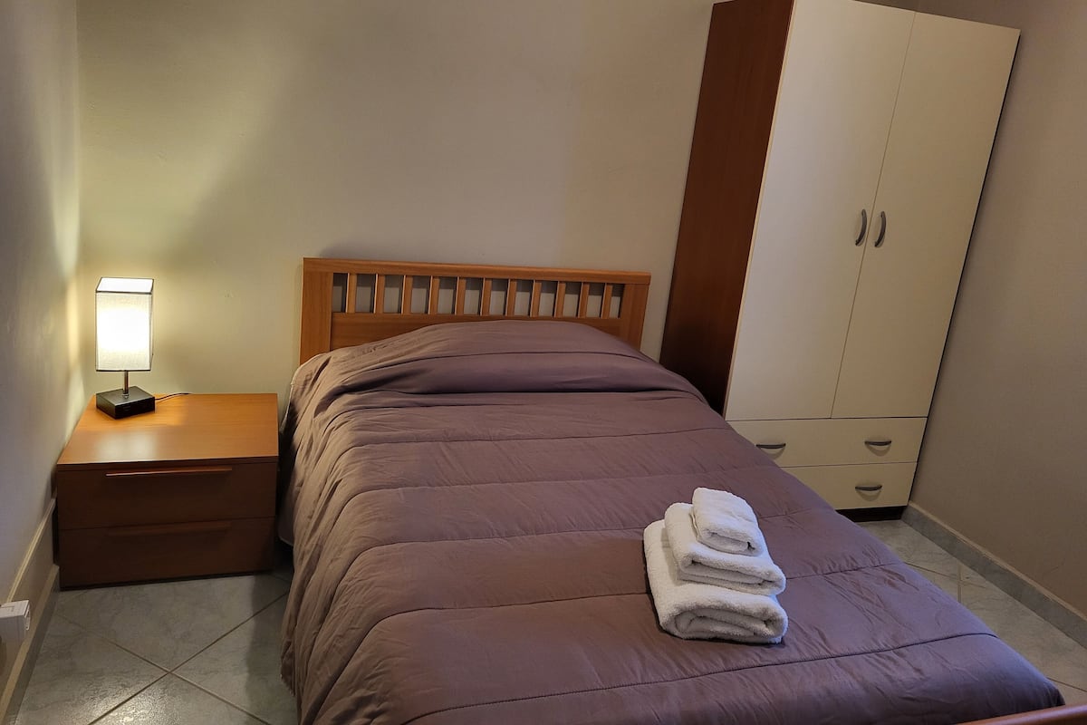 Ein Schlafzimmer mit einem Bett, einem Nachttisch, einer Lampe, einem Kleiderschrank und einem Stapel Handtücher.