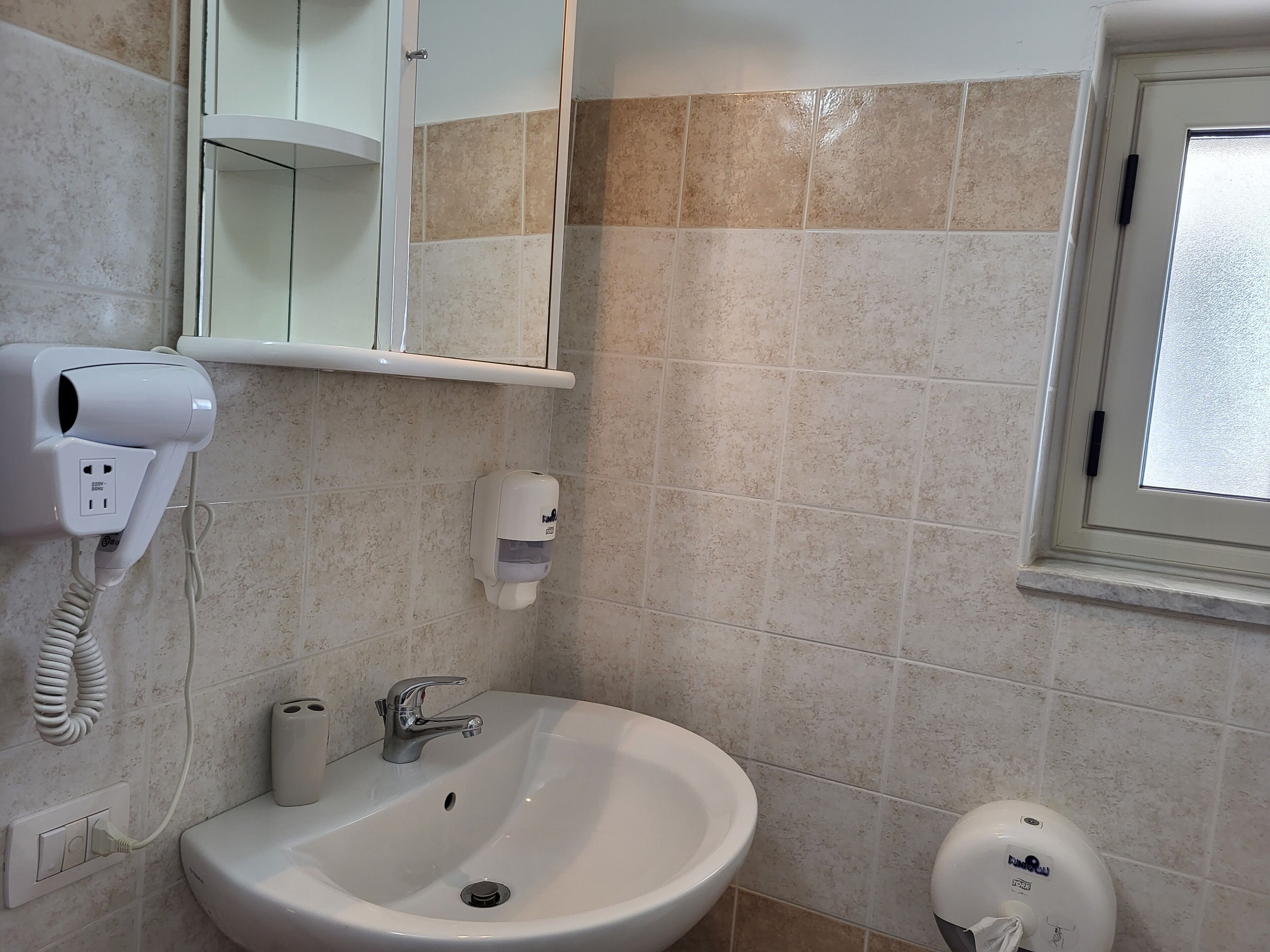 Une salle de bain avec un lavabo blanc, un sèche-cheveux, un distributeur de savon et une fenêtre.