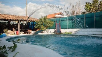Uma piscina com uma fonte de água, um escorregador vermelho e uma cerca verde.