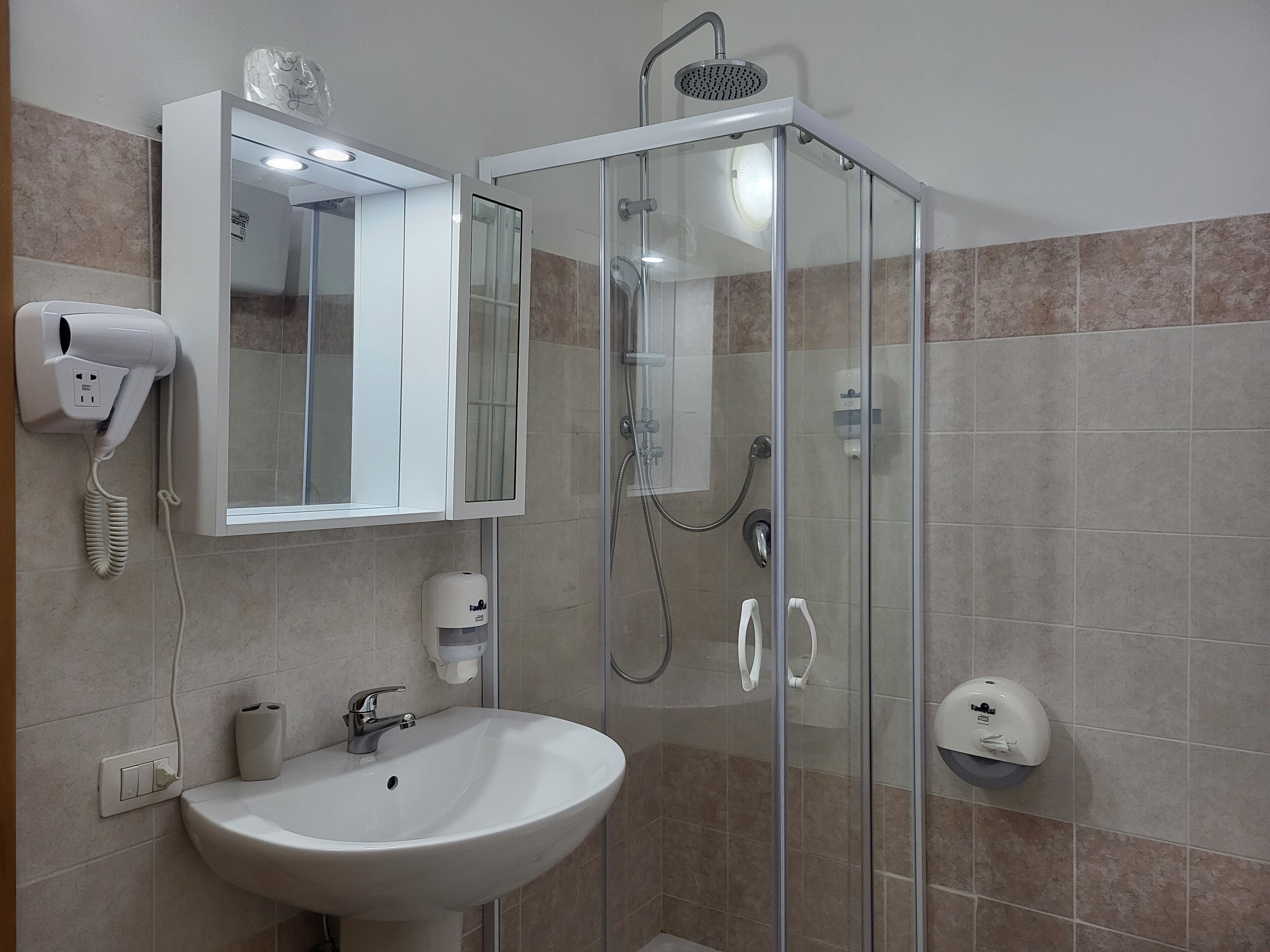Ein Badezimmer mit Duschkabine, Waschbecken in Weiß, einem Spiegel und einem an der Wand befestigten Föhn.