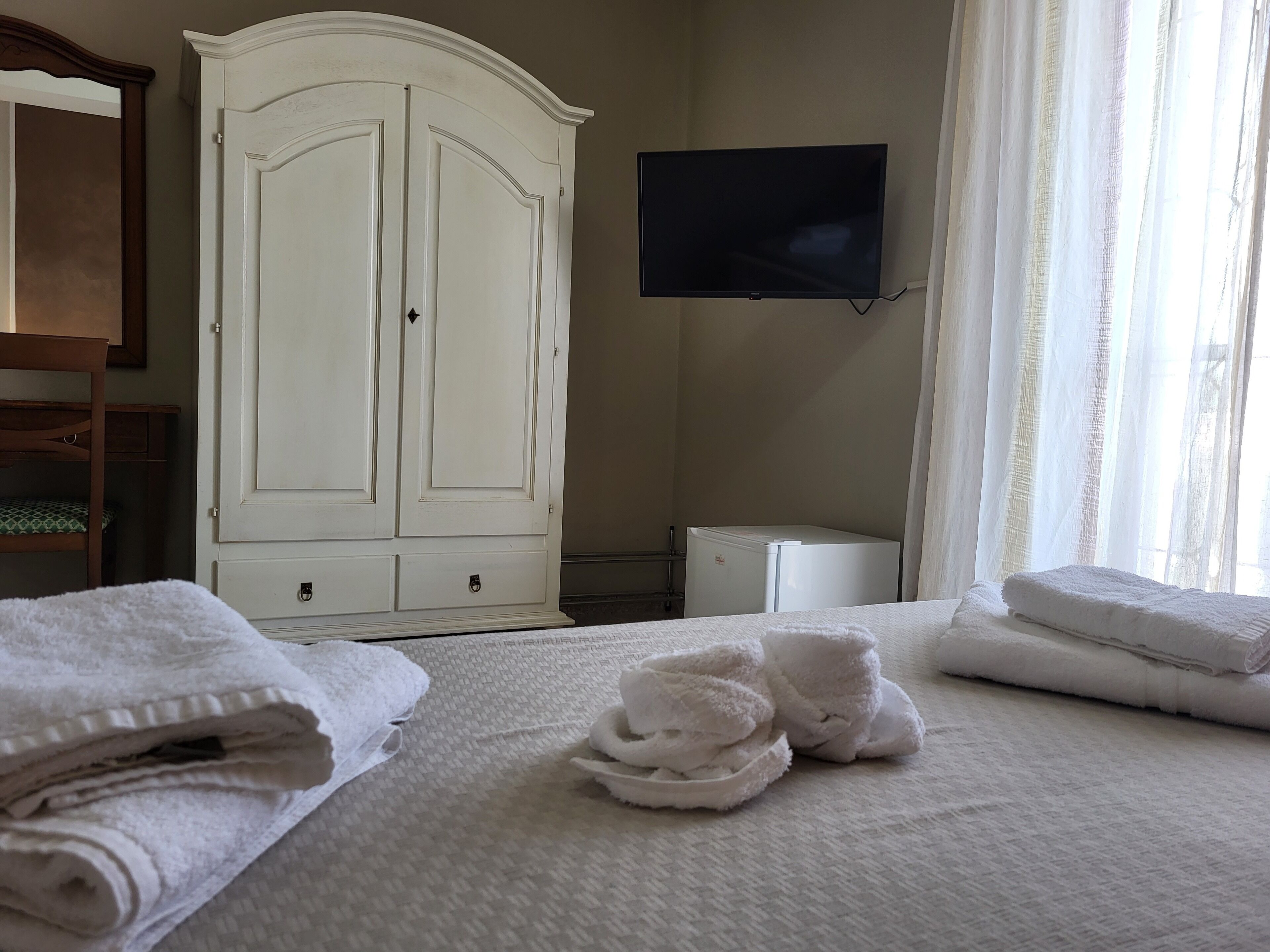 Une chambre d’hôtel avec une grande armoire blanche, un téléviseur à écran plat fixé au mur, un petit réfrigérateur blanc et des serviettes soigneusement pliées sur le lit.