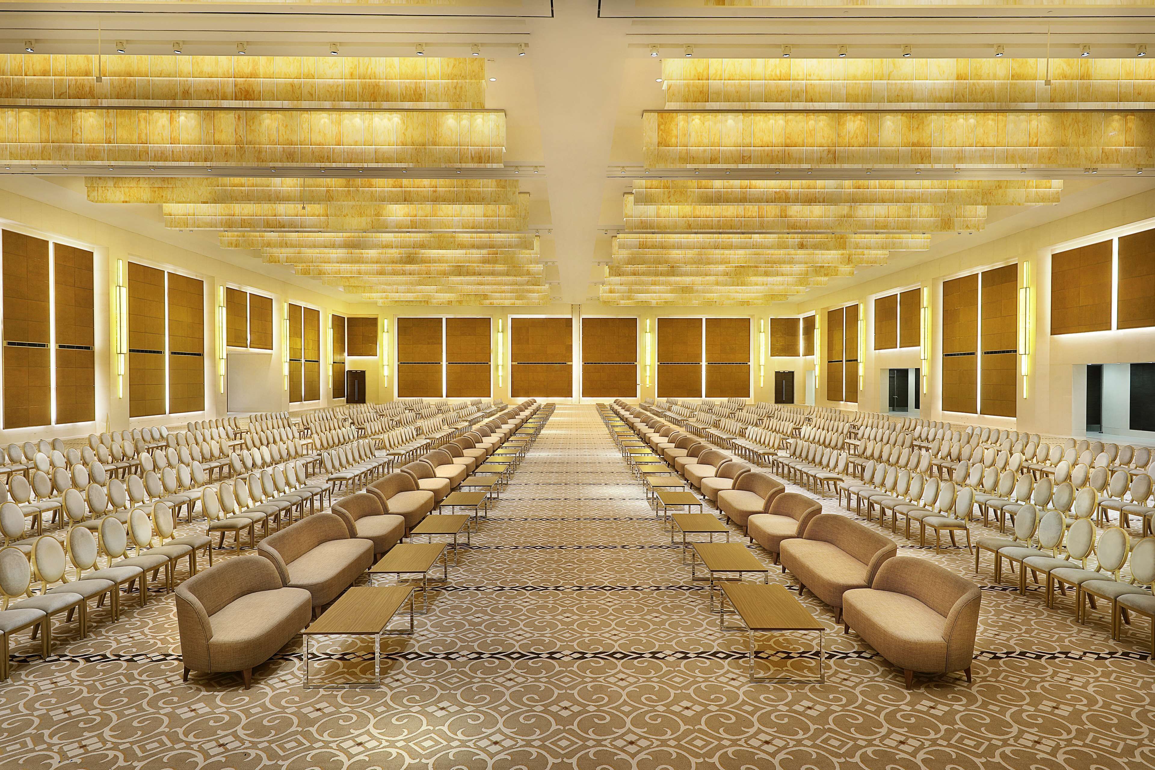 A large conference room with rows of chairs and sofas, a patterned carpet, and a ceiling with horizontal lighting fixtures.