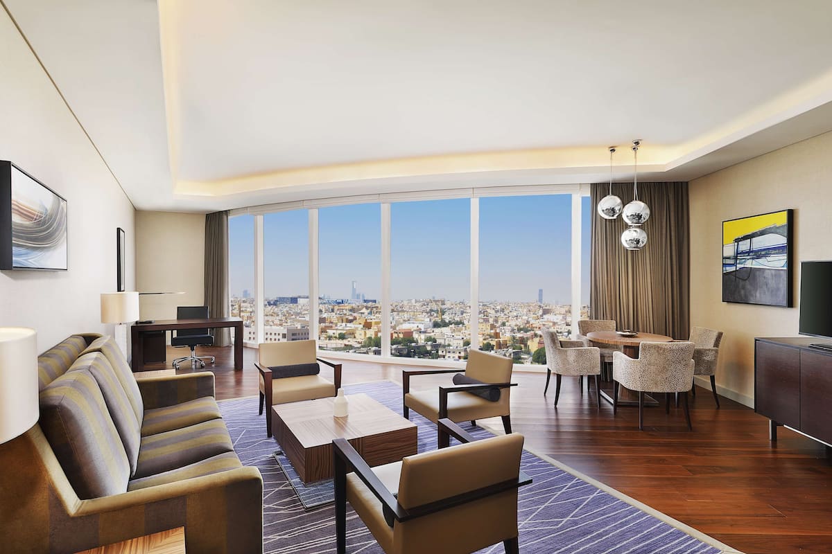 Una habitación de hotel moderna con un ventanal grande, una zona de comedor y un área de descanso con sofá y sillones.