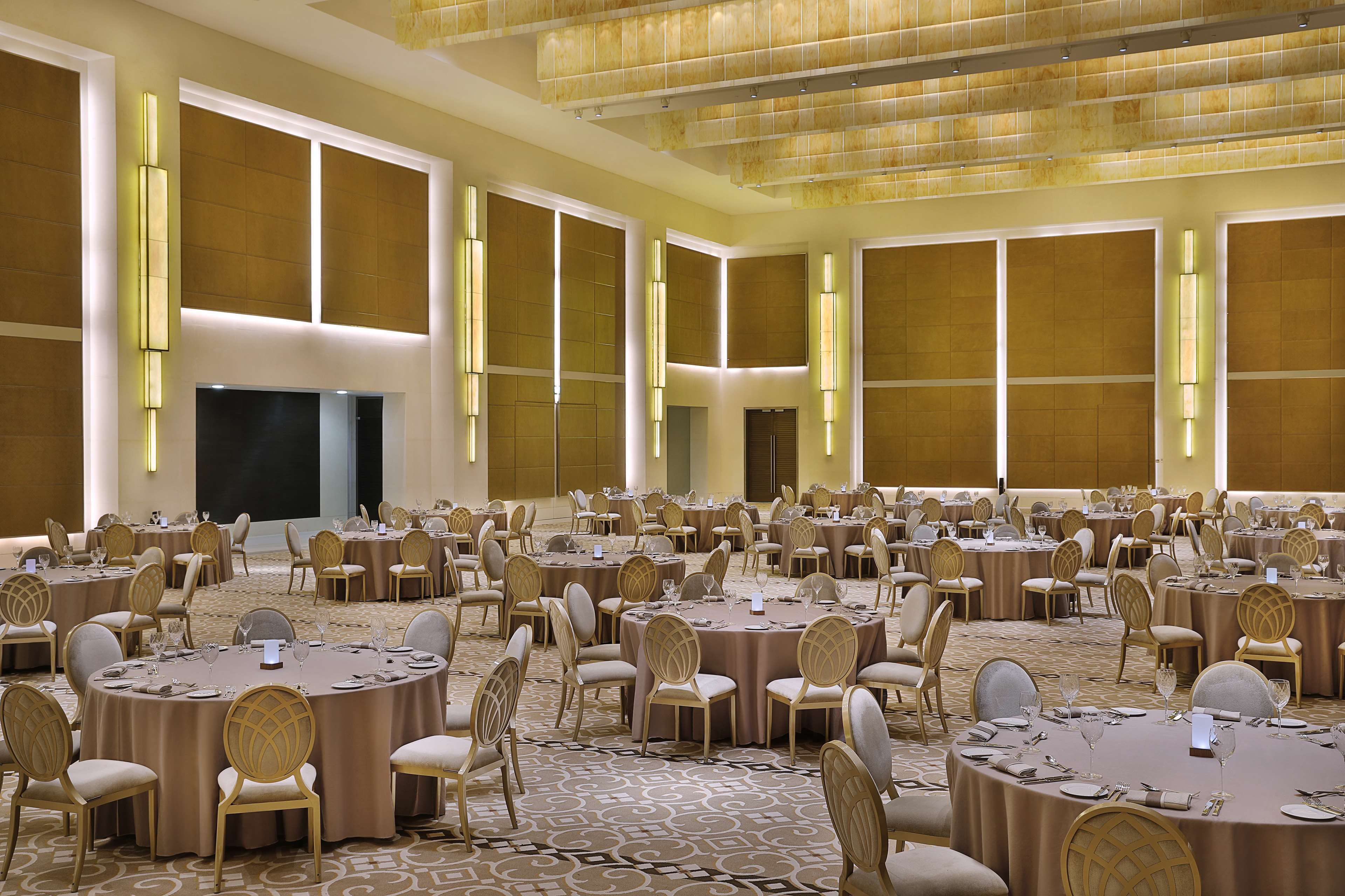 A large banquet hall with round tables and chairs, a carpeted floor, and a high ceiling with recessed lighting.