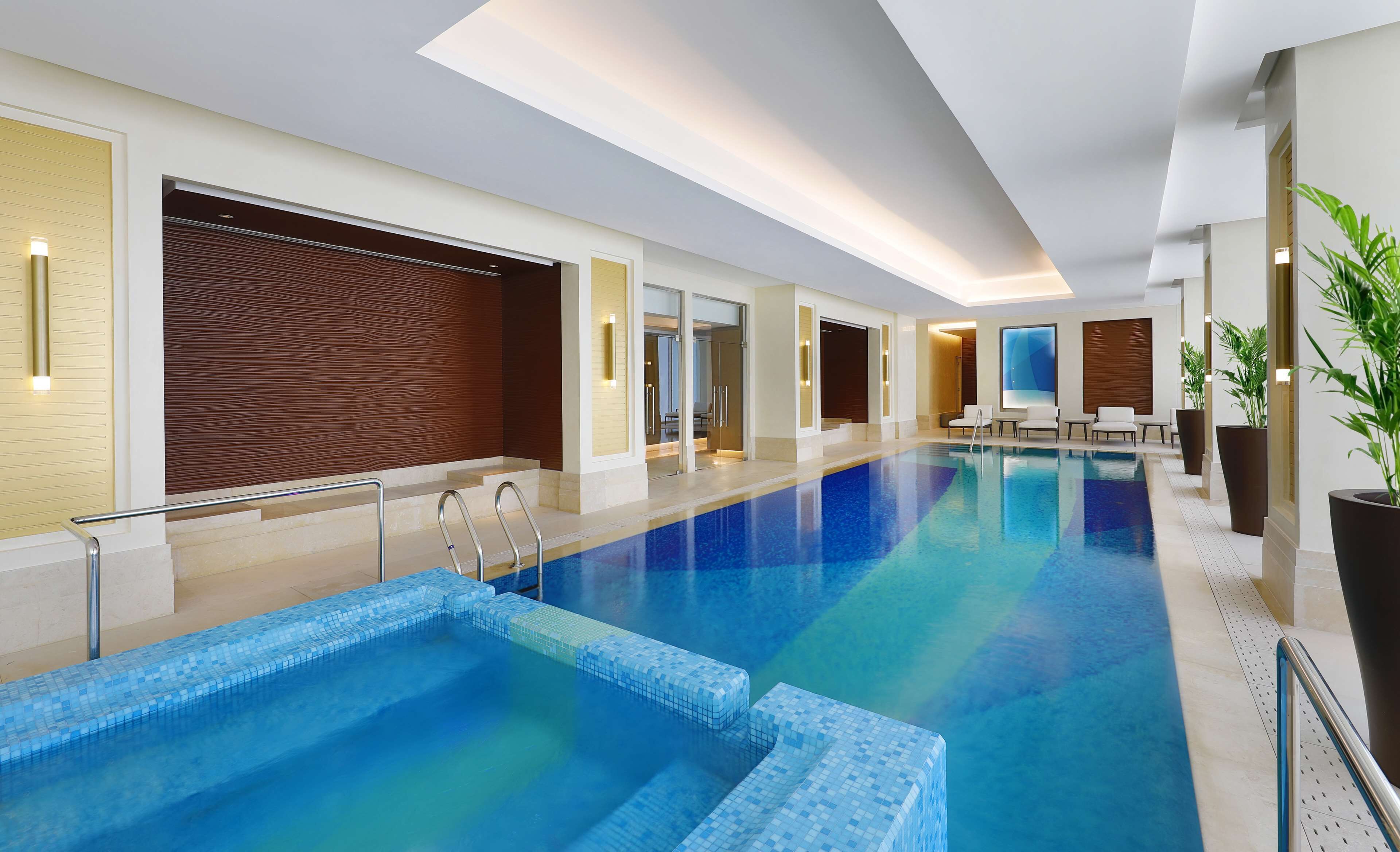 A modern indoor swimming pool with clear blue water, a tiled deck, and a seating area with chairs and tables.