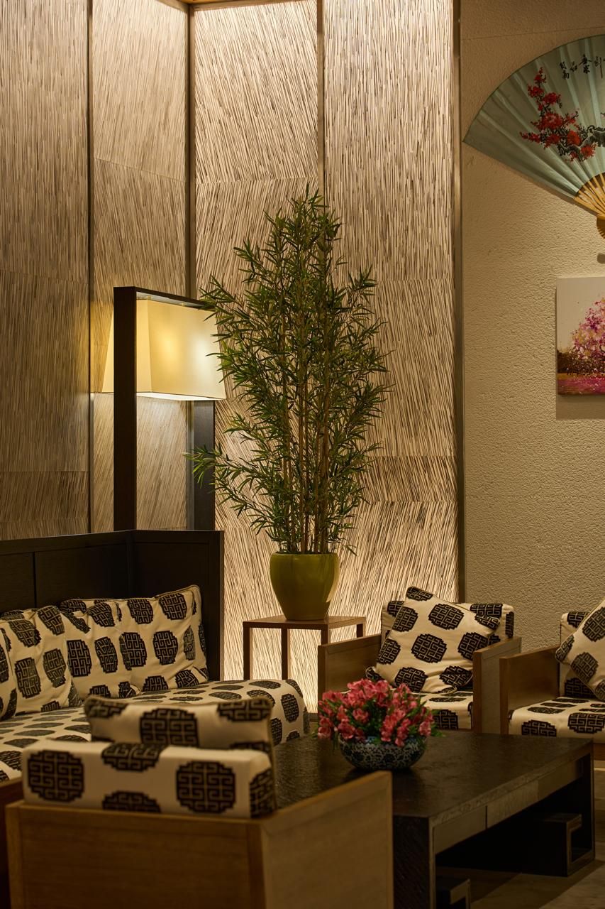 Una habitación con decoración de estilo japonés tradicional, que incluye una mesa baja de madera, cojines estampados, una planta de bambú en maceta y un ventilador de pared.