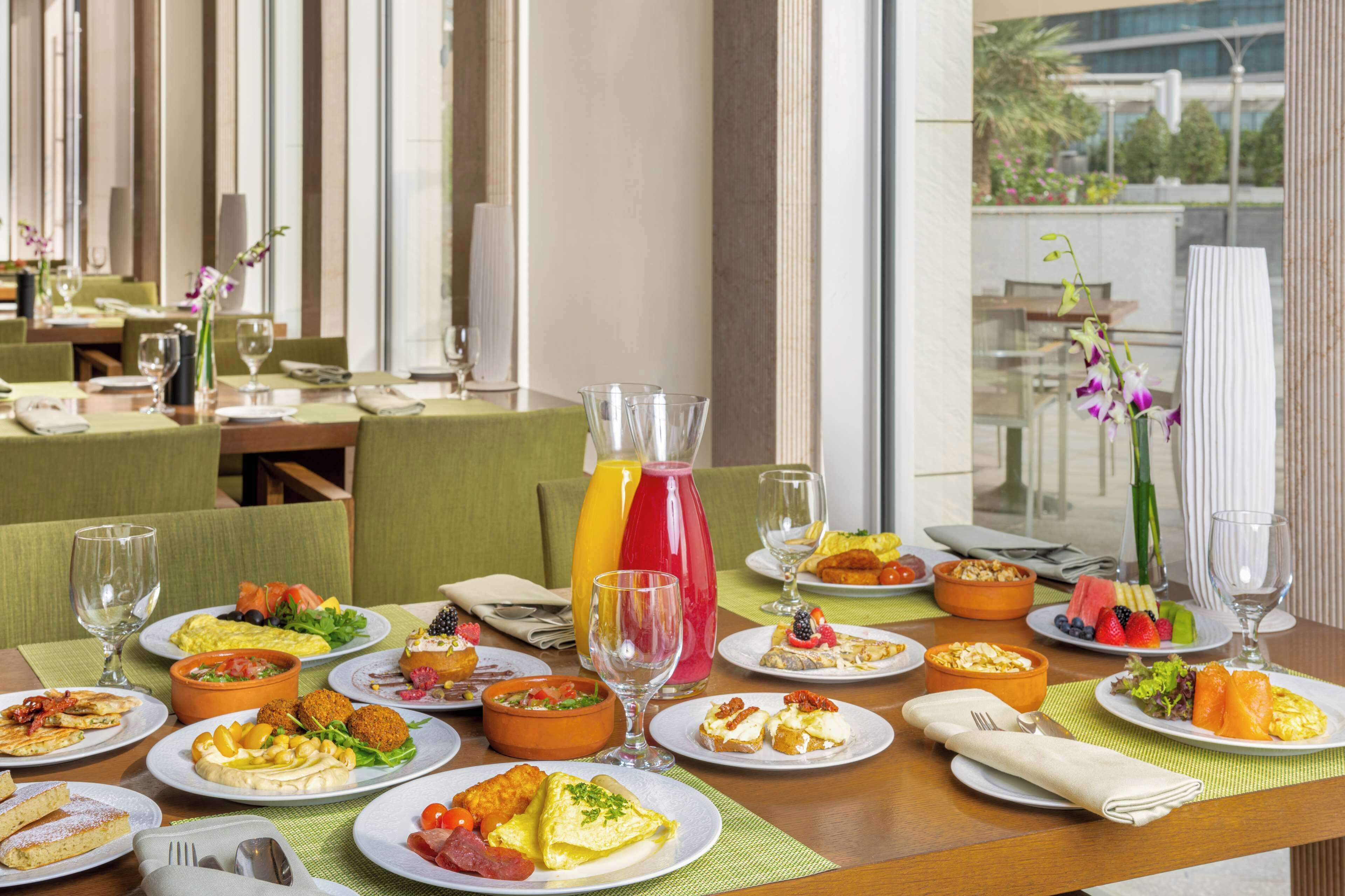 Ein Esszimmer mit einem gedeckten Tisch, auf dem verschiedene Frühstücksleckereien wie Eier, Früchte und Gebäck ausliegen, mit großen Fenstern und viel Licht.