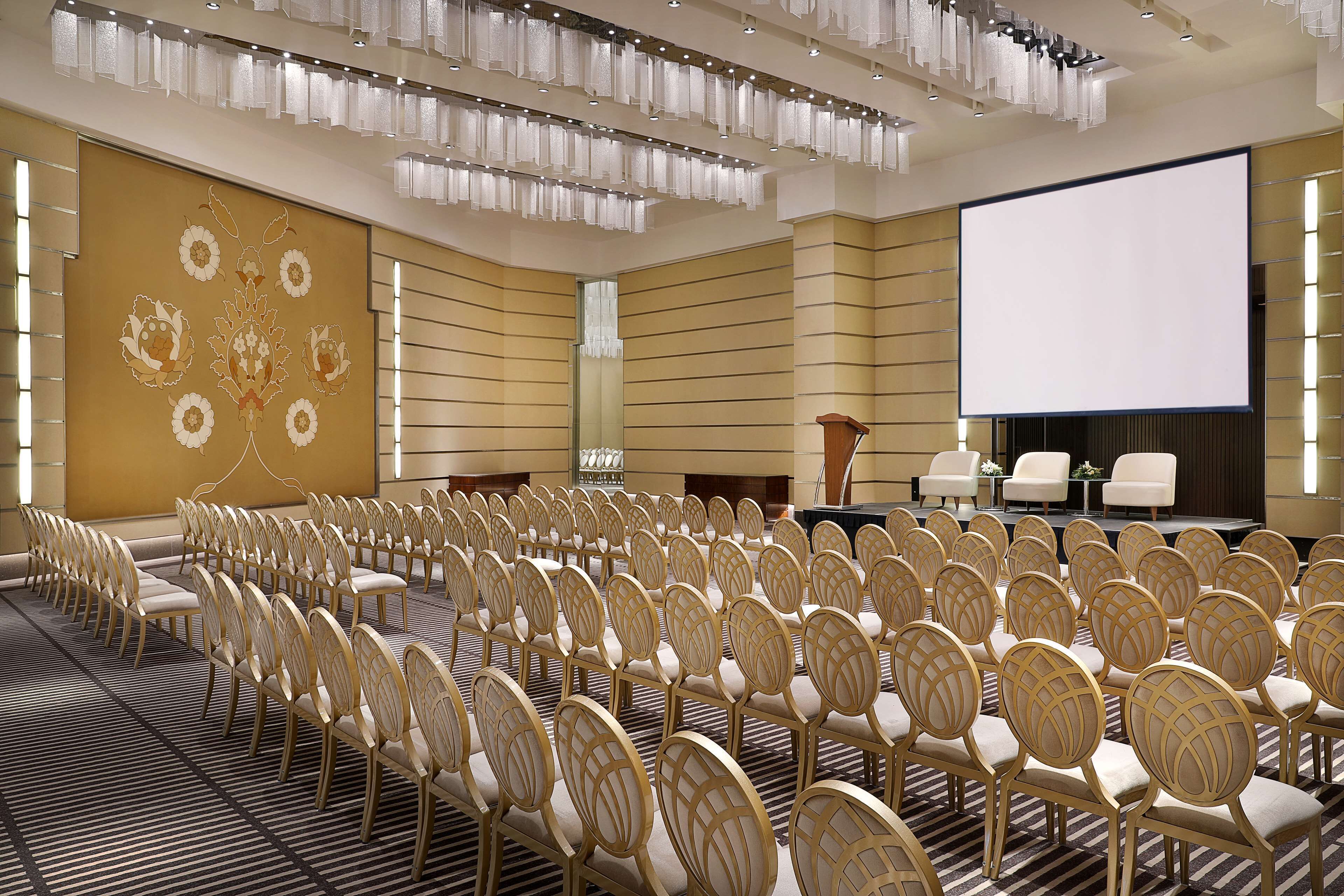 Un salón de conferencias con filas de sillas, una pantalla grande y un escenario con un atril.