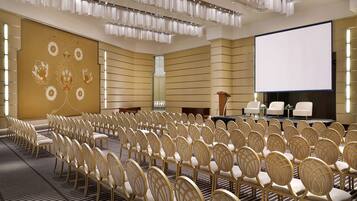 Un salón de conferencias con filas de sillas, una pantalla grande y un escenario con un atril.