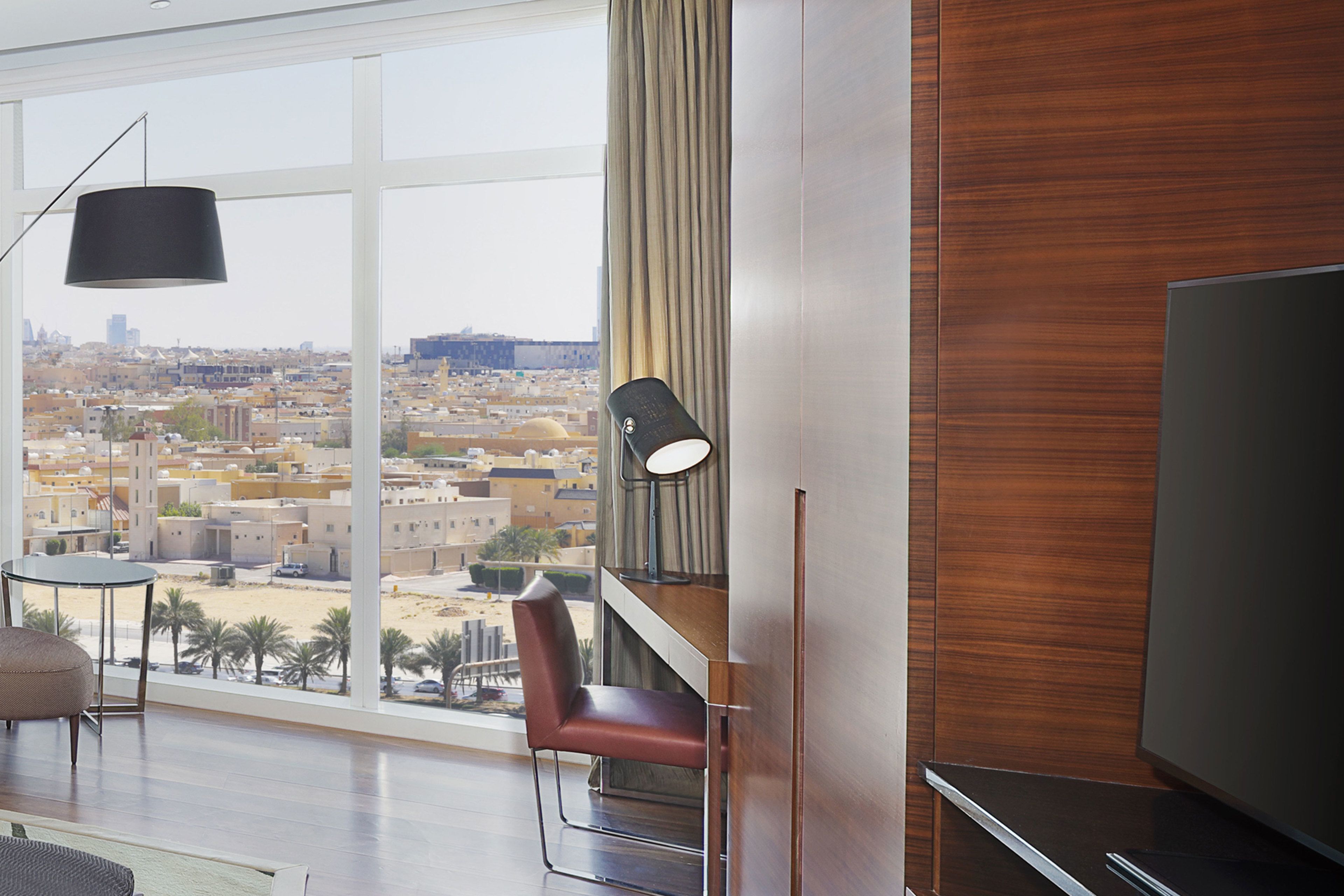 A modern hotel room with a large window offering a city view, a flat-screen TV, a comfortable chair, and a stylish lamp.