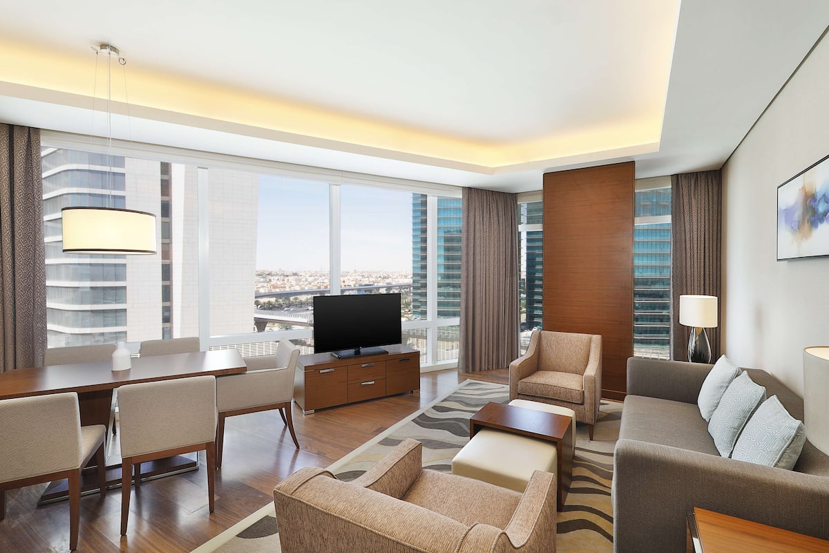 Una habitación de hotel moderna con un ventanal grande, un televisor de pantalla plana, una zona de comedor y un sofá.