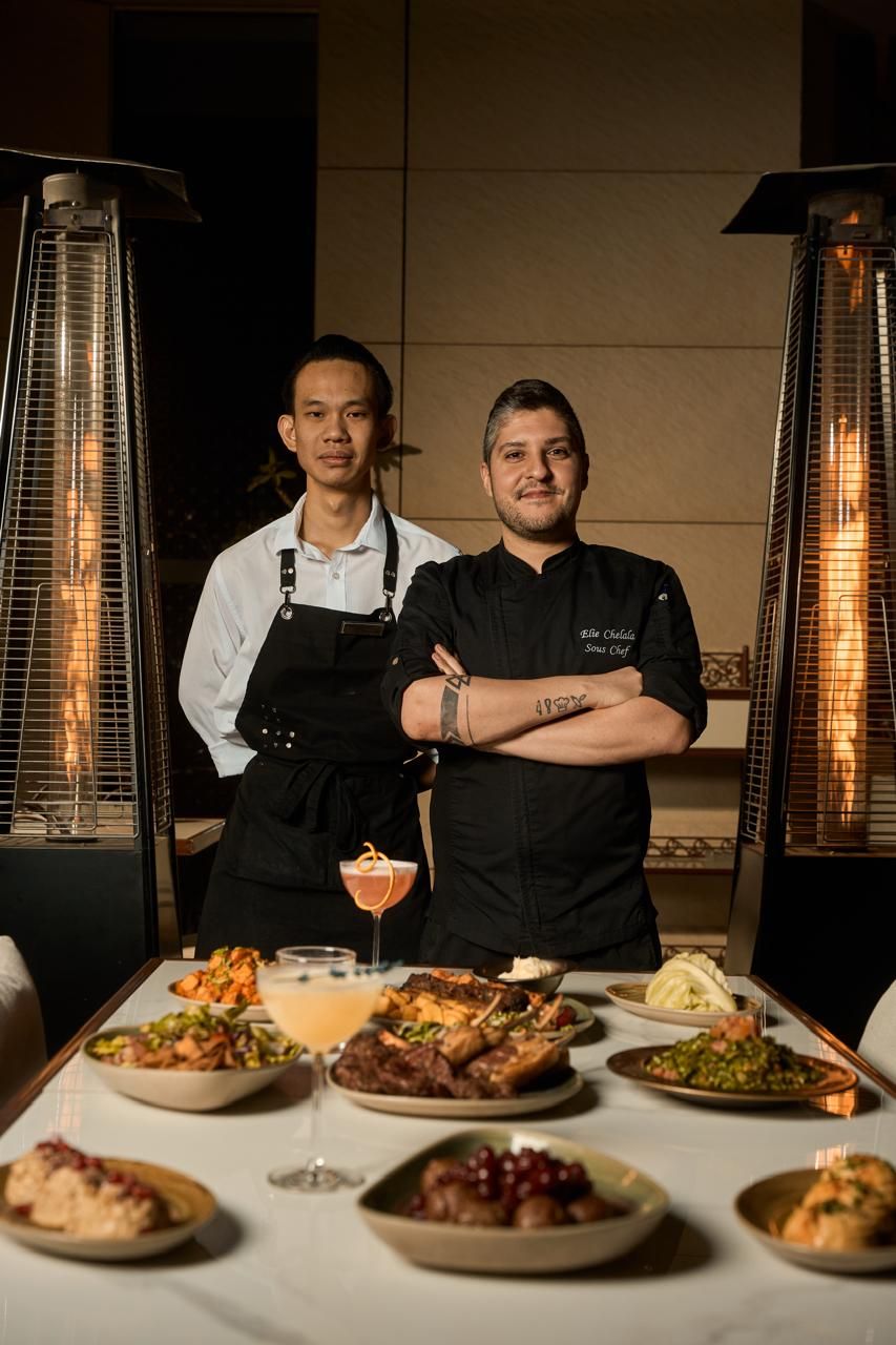 Two chefs standing behind a table with various dishes and a drink.
