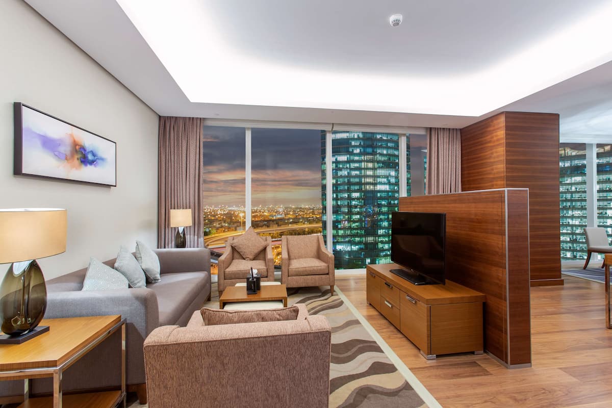Una habitación de hotel moderna con un ventanal grande que ofrece vistas a la ciudad, un televisor de pantalla plana, un sofá, una mesita de café y un armario de madera.