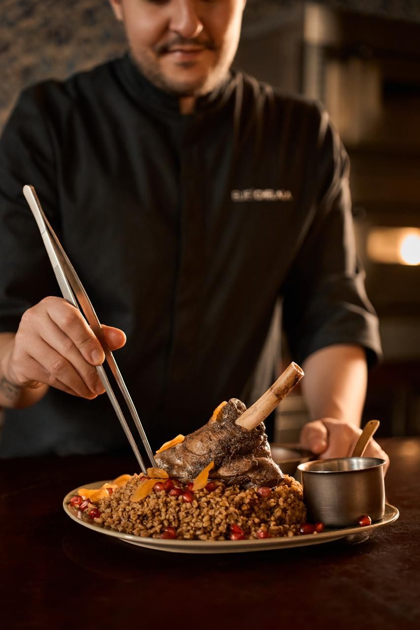 A chef holding chopsticks over a plate of food, including a lamb shank and grains, with a small metal bowl on the side.