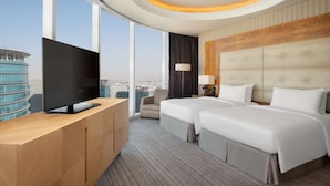Habitación de hotel con televisor grande, dos camas, escritorio y vista a la ciudad.