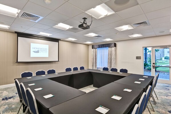 A conference room with a long table, chairs, a projector screen displaying "Homewood Suites by Hilton," and a door leading outside.