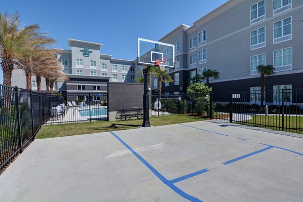 A basketball court with a net, palm trees, and a pool area in front of a hotel building. - Homewood Suites by Hilton New Orleans Westbank (Gretna)