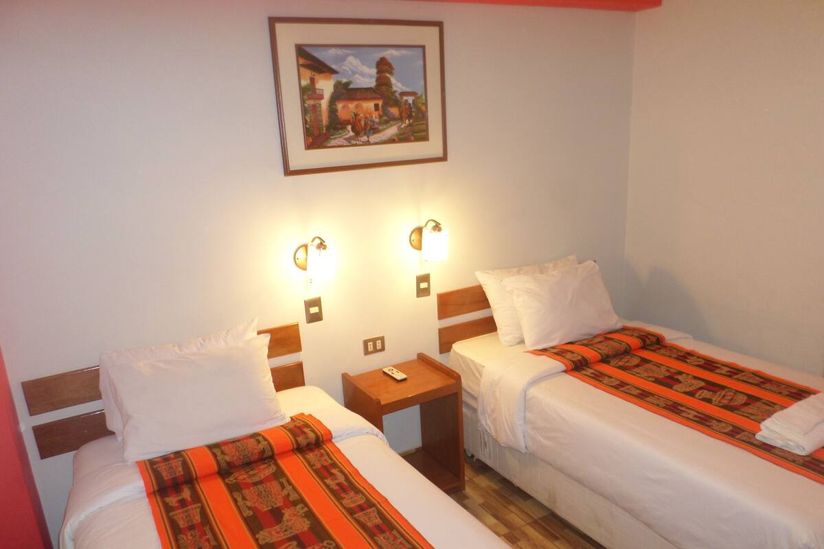 A hotel room with two beds, each with white linens and a patterned blanket. There are two bedside tables with lamps, a framed picture on the wall, and a wooden floor.