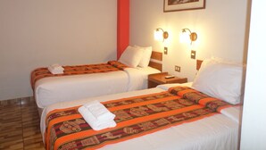 A hotel room with two beds, each with a patterned blanket and pillows, a nightstand with a remote control, and a painting on the wall. - Hatun Pueblo Inn (Machu Picchu)