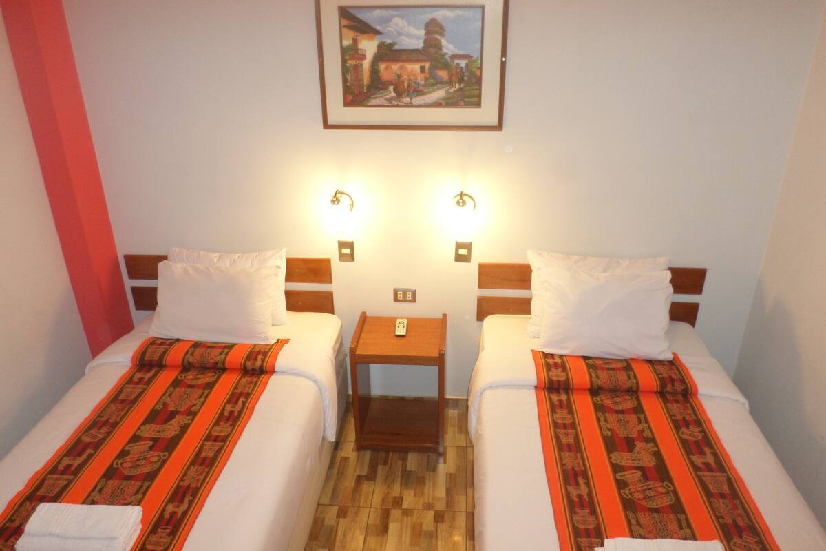 A hotel room with two beds, each with a patterned bedspread and white pillows. There are two bedside tables with lamps, a small nightstand, and a framed picture on the wall.