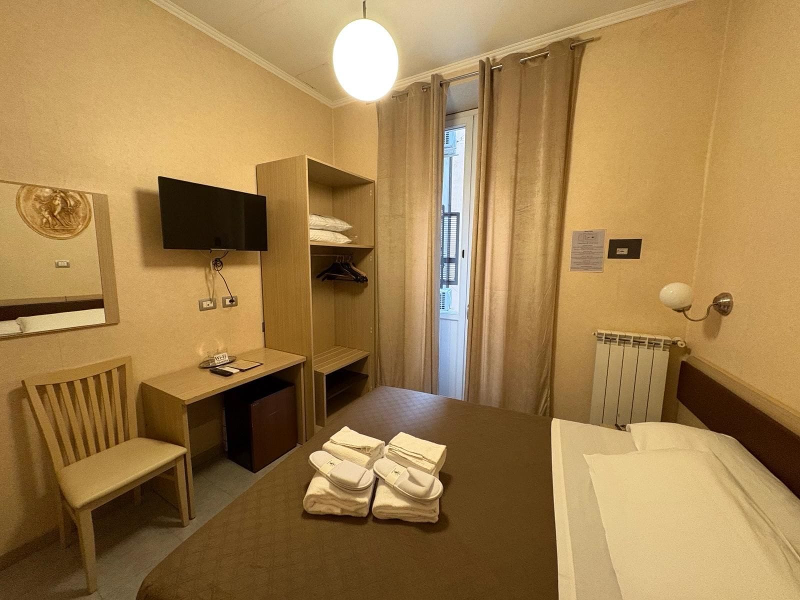 A hotel room with a bed, a desk, a chair, a TV, a wardrobe, and a radiator.