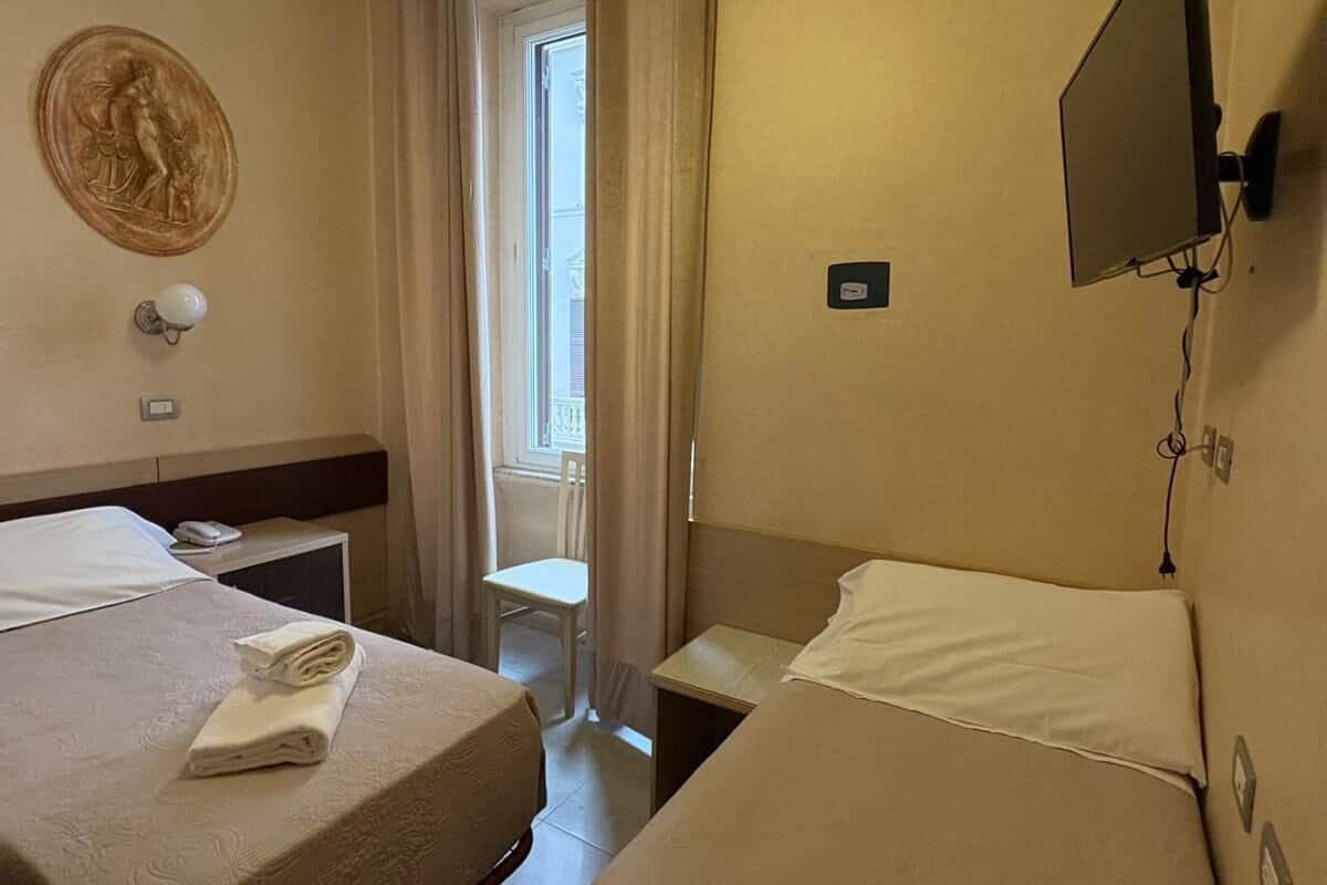 Habitación de hotel con dos camas, un televisor de pantalla plana montado en la pared, una ventana con cortinas y una lámpara de pared.