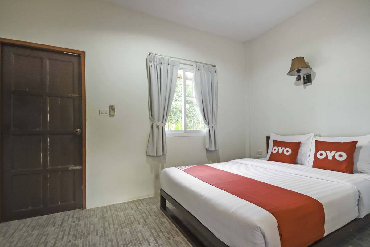 A hotel room with a bed, a window with curtains, a wall-mounted lamp, and a door.