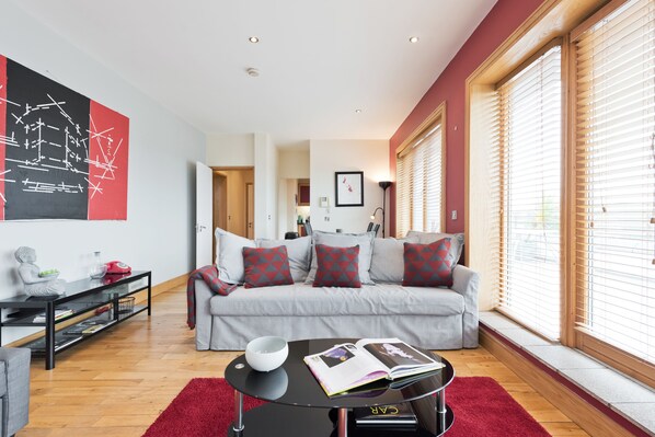 Una sala de estar moderna con un sofá gris, cojines estampados en rojo y negro, una mesa de centro negra con un cuenco y una revista, y un cuadro abstracto grande en la pared.