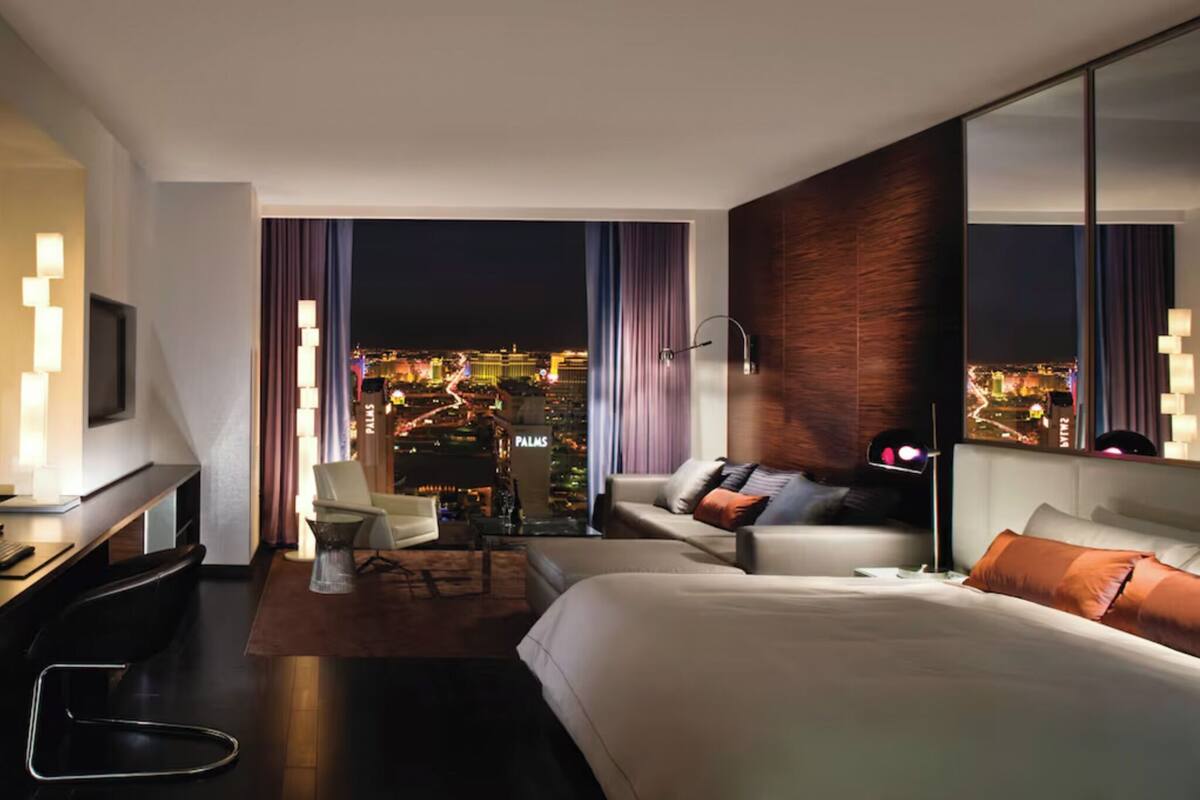 Une chambre d’hôtel moderne avec un grand lit, un bureau, une chaise et une télévision. La chambre offre une vue sur la ville, et les lumières sont visibles à travers la fenêtre.